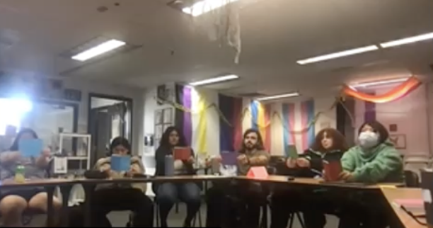 A group of people sitting around a table, each holding up a colorful sheet of paper, in a room decorated with rainbow flags.